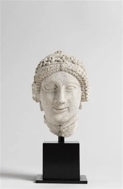 Tête de femme coiffée d'un kékryphale : chignon rejeté en arrière, bandeau, diadème à 7 rosettes en relief, cordon autour du cou (pendeloque non conservée), couvre-oreilles en forme de peignes à 4 dents, boucles d'oreilles circulaires à bouton central, restes de pigments rouges au niveau des boucles d'oreilles